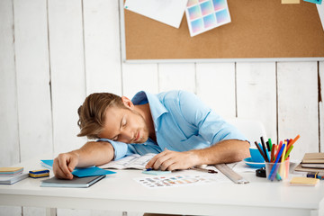 Young handsome sleepy tired businessman sleeping at table on papers and notepad. White modern office interior background.