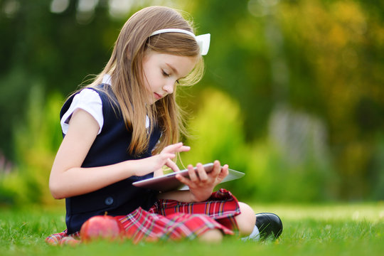 Adorable little girl using computer tablet while sitting on a grass on summer day