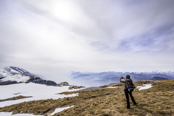 Fototapeta premium Girl taking a picture of mountain landscape in the middle of a field with some snow at Monte Altissimo, San Giacomo, Trentino region, Italy