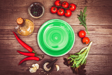 Fresh tomatoes, chili pepper and other spices and herbs around modern green plate in the center of wooden table and cloth napkin. Top view. Blank place for your text. Close-up.