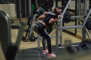 Young strong woman doing squats at the gym in front of a mirror with a barbell on her back
