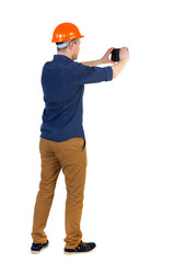 Backview of business man in construction helmet stands and enjoys tablet or using a mobile phone. Standing young guy. Rear view people collection.  backside view of person.  Isolated over white