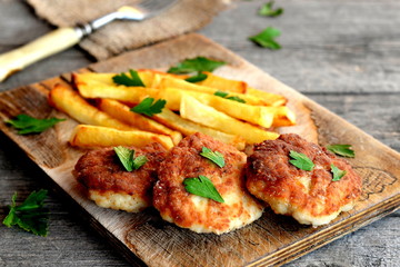 Meat cutlets served with deep fried potatoes. Turkey breast burgers with vegetables on cutting board. Fork, sprig of fresh parsley on an old wooden background. Lunch or dinner idea. Closeup