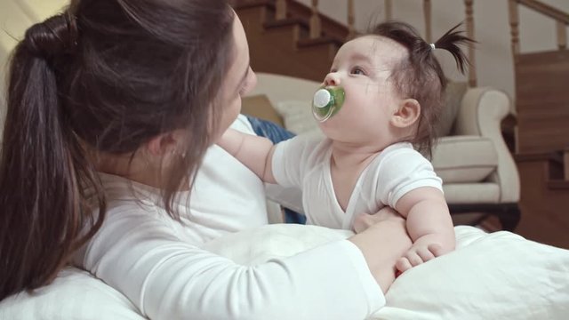 Woman lying on bed, holding and kissing adorable baby girl while she sucking a soother