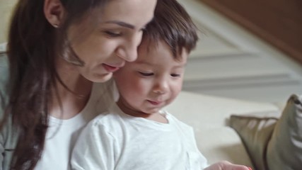 Cute boy sitting on his knees of his mother while she showing him something on tablet screen
