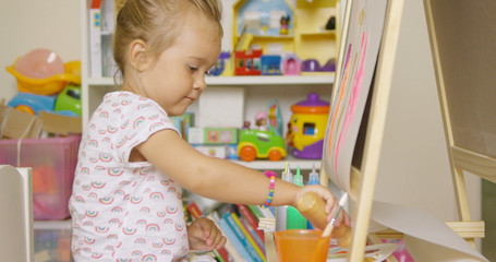 Little girl having fun painting with water color paints at an easel in her playroom selecting paint from a bottle for her palette