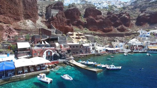 Aerial footage of Oia, Santorini - Famous white houses and blue domes on the edge of the cliff and blue lagoon.