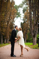Stylish bride in beige dress kisses a groom on the path in park