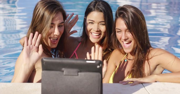 Three vivacious smiling women taking a selfie on a tablet computer at the side of a swimming pool laughing and joking as they pose for the camera.