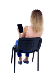 back view of woman sitting on chair and looks at the screen of the tablet.  Rear view people collection.  backside view of person.  Isolated over white background. Blonde in blue trousers sitting on