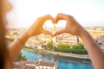 Female hands making heart shape on Verona cityscape background. Verona is famous city of love in the north of Italy. - obrazy, fototapety, plakaty