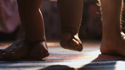 Close-up of baby boy walking his first steps with dad behind, at sunny room