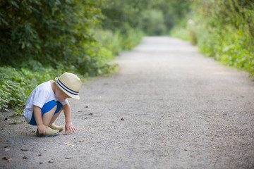 Cute little toddler boy in straw hat palying on the countryside road on a sunny summer day. Lifestyle concept