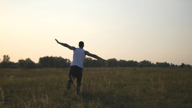 Young man with raising hands running in field and enjoying freedom. Carefree guy running on grass field at summertime. Landscape background. Relaxation on nature. Rear back view