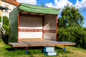 Makeshift stage in a rural garden. Stage is made of garage jacks supporting a wooden floor and scaffolding railings covered with a green tarp as roof and walls. - 118058949