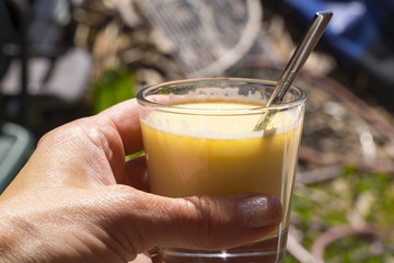 First person perspective on hand holding curcuma milk in glass with metal spoon