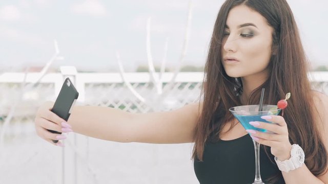 Young girl posing for selfie with goblet of blue cocktail on the beach. Slowly