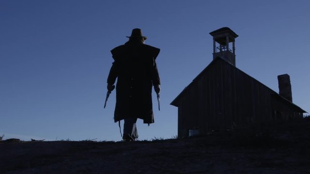 Silhouette of a cowboy with a gun in each hand, walking to a building. 