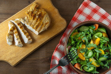 Chicken fillet, grilled, with a large portion of salad with fresh vegetables and herbs on a wooden table with a red cloth into a cell. top view