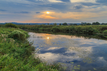 The sun sets in the field across the river