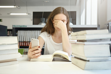 Tired female student of school of economics sitting at library rubbing her eyes, bored with reading audit manual, checking time using her smart phone application, surrounded by stacks of books