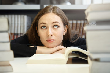 Cute brunette student girl in black jacket studying and reading textbook or manual in university library but having hard time understanding material, rolling her eyes, looking bored and confused