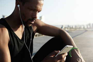 Nice photo of young and handsome athlete choosing musical track for running on digital device. Lonely African American man taking a break from his workout and enjoying beautiful song in headphones.
