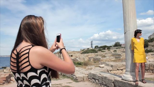 Happy women taking photo girl friend with smartphone on the ruins of the ancient city background