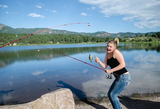 Young excited girl fishing with thumbs up with a caught fish on hook on a beautiful sunny day with greenery, mountains, trees and blue cloudy day at Monument Lake, CO.