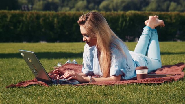Attractive barefoot young woman relaxing with her laptop on the grass lying on a rug in the garden typing or surfing the internet