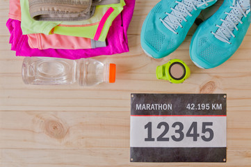 Top view of outfit for runner on wooden background: bib number, bottle of water, gps watch, running shoes, running waist bag, shorts, shirt, and sport bra. Horizontal orientation.