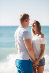 Young happy couple in love,blond man and woman spend time together on the beach on the shore of the blue ocean,standing on the warm sand holding hands on a background of calm sea
