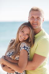 Happy young couple blond man and woman spend time together on the beach on the shore of the blue ocean,standing on the sand in an embrace on a background of calm sea and clear sky