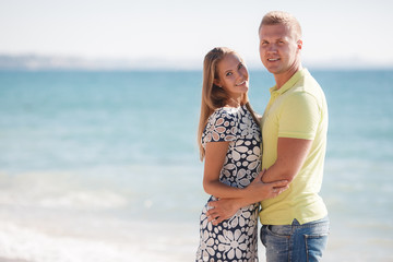 Happy young couple blond man and woman spend time together on the beach on the shore of the blue ocean,standing on the sand in an embrace on a background of calm sea and clear sky