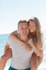 Happy loving couple,young blond woman having fun on the beach near the blue ocean,a girl is sitting on the back of a guy hugging his neck the girl wears a ring on his right hand,he wears a gold chain