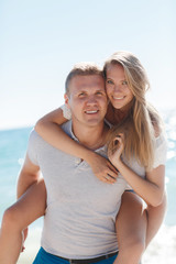Obraz premium Happy loving couple,young blond woman having fun on the beach near the blue ocean,a girl is sitting on the back of a guy hugging his neck the girl wears a ring on his right hand,he wears a gold chain