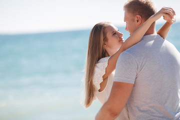 Fototapeta premium Happy young couple blond man and woman spend time together on the beach on the shore of the blue ocean,standing on the sand in an embrace on a background of calm sea and clear sky