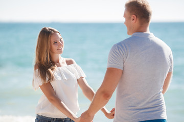 Young happy couple in love,blond man and woman spend time together on the beach on the shore of the blue ocean,standing on the warm sand holding hands on a background of calm sea
