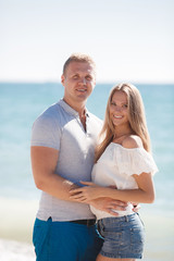 Happy young couple blond man and woman spend time together on the beach on the shore of the blue ocean,standing on the sand in an embrace on a background of calm sea and clear sky