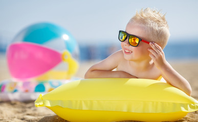 A little boy with a short haircut and blonde hair,cute smile, dark sunglasses,lying on a yellow air...