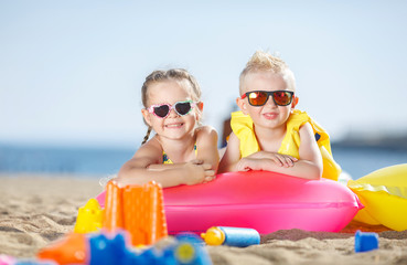 Little girl with pigtails and a little boy with short hair,wearing dark sun glasses, spend time together on the sandy beach,lie side by side on air mattresses for swimming on the blue ocean