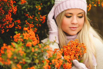 Naklejka premium young woman with long blonde straight hair,wearing a light sweater,pink scarf and gloves,on his head wearing a pink and white knitted cap,spends time outdoors in the Park near bushes of ripe red Rowan
