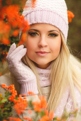 young woman with long blonde straight hair,wearing a light sweater,pink scarf and gloves,on his head wearing a pink and white knitted cap,spends time outdoors in the Park near bushes of ripe red Rowan