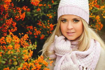 young woman with long blonde straight hair,wearing a light sweater,pink scarf and gloves,on his head wearing a pink and white knitted cap,spends time outdoors in the Park near bushes of ripe red Rowan