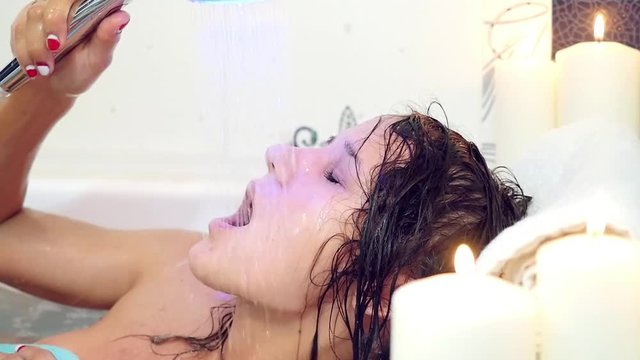 beautiful young brunette woman taking a relaxing bath with foam and pouring water on herself. Burning candles at backround
