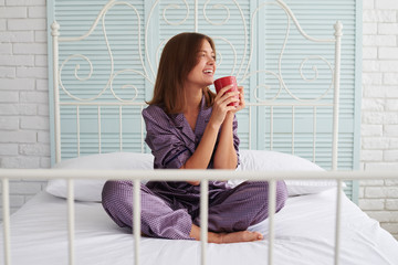 Brown-haired lady in pajamas drinking her morning coffee on bed