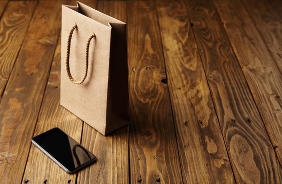 Light brown craft paper bag reflecting in a spotless black smartphone lying next to it on a handcrafted wooden table