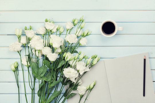 Morning coffee cup, empty paper list, pencil, and bouquet of white flowers eustoma on blue rustic table overhead view. Woman working desk. Cozy breakfast. Flat lay styling.