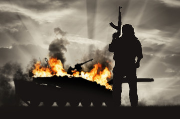 Silhouette of terrorist holding rifle against burning tank - obrazy, fototapety, plakaty
