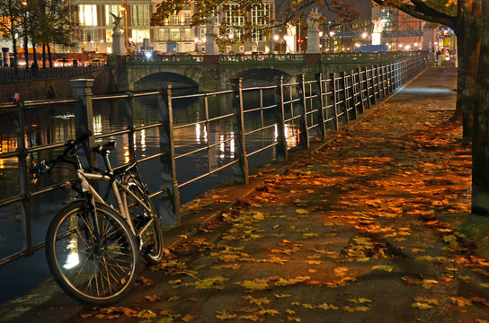 Bicycle resting against a fence along a city canal at night, during autumn in Berlin, Germany.